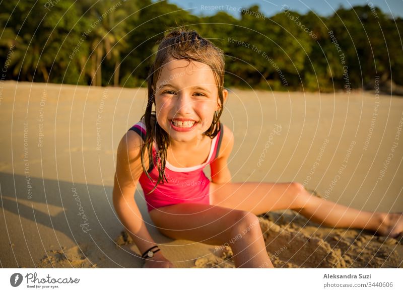 Cute happy little girl playing with sand on the beach in swimming suit, drawing a heart and writing. Beautiful summer sunset, sea, coconut palms, picturesque exotic landscape. Phuket, Thailand