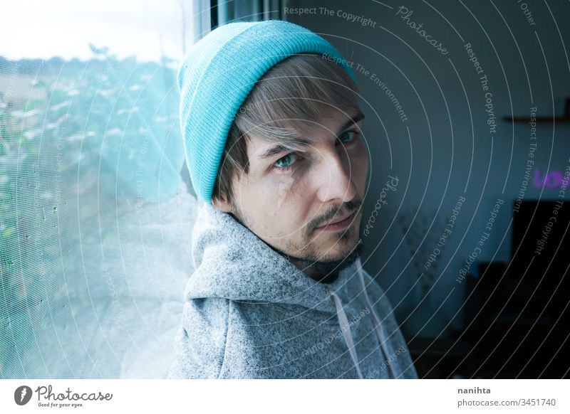 Young man alone in his home near a window male portrait isolated locked quarantine lonely self attractive handsome guy casual wear hairstyle blue eyes lenses