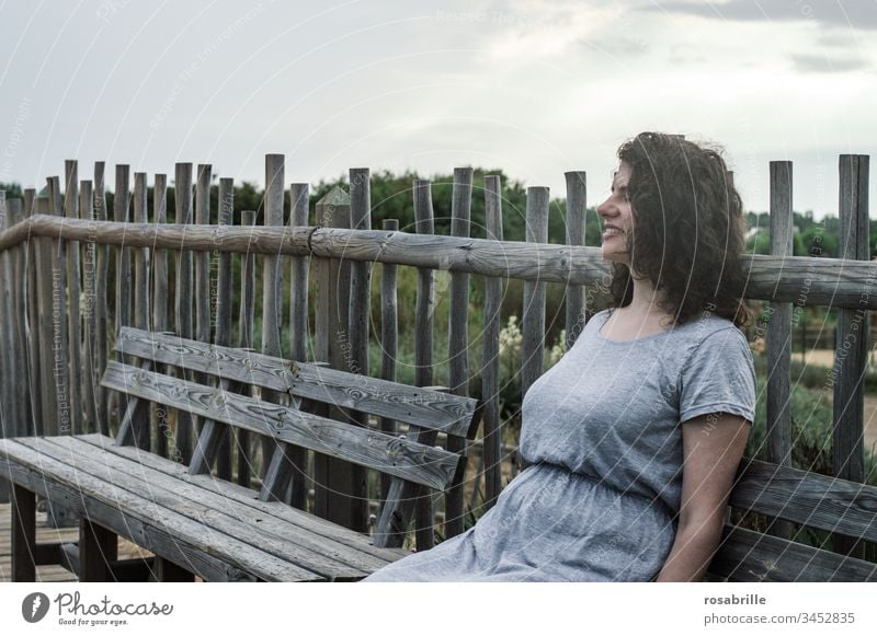 young pretty brunette woman with curly hair sits on a wooden bench in front of a board fence in the dunes near the beach and enjoys the holiday youthful