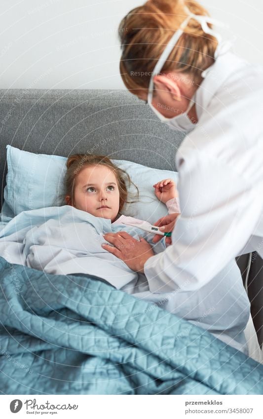 Doctor visiting little patient at home. Measuring the temperature of sick girl lying in bed. Woman wearing uniform and face mask. Medical treatment child virus