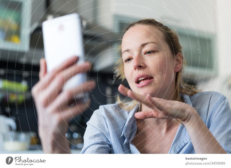 Young smiling cheerful pleased woman indoors at home kitchen using social media apps on mobile phone for chatting and stying connected with her loved ones. Stay at home, social distancing lifestyle.