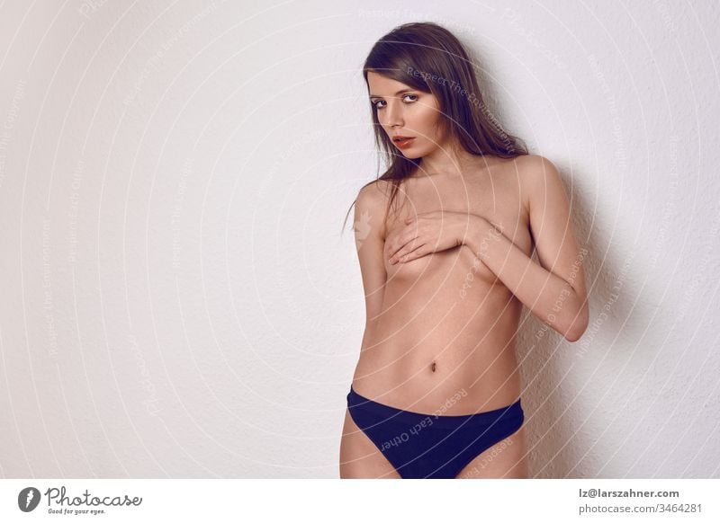 Topless sexy young brunette woman in blue panties, covering her breast with one hand, stands on a light background in the studio and looks into the camera with a seductive look. Front portrait with copy room