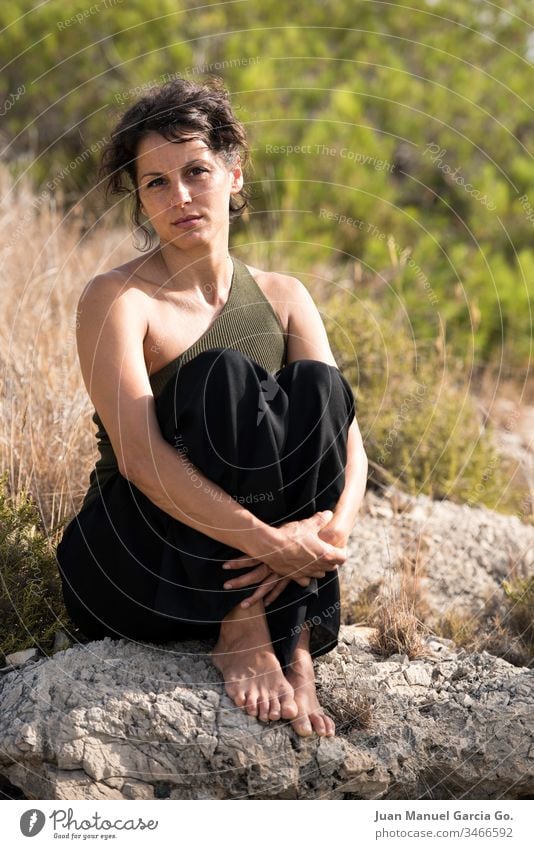 A vertical shot of a beautiful female Latina sitting on a rock by the bush woman beauty white girl young nature pretty portrait adult background attractive