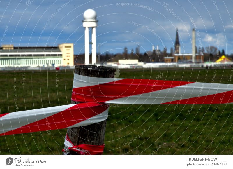 Corona Thoughts | The Tempelhofer Feld leisure area is marked with red and white barrier tape and in the background, against a dark cloudy sky, the striking airport building glows in the sunlight