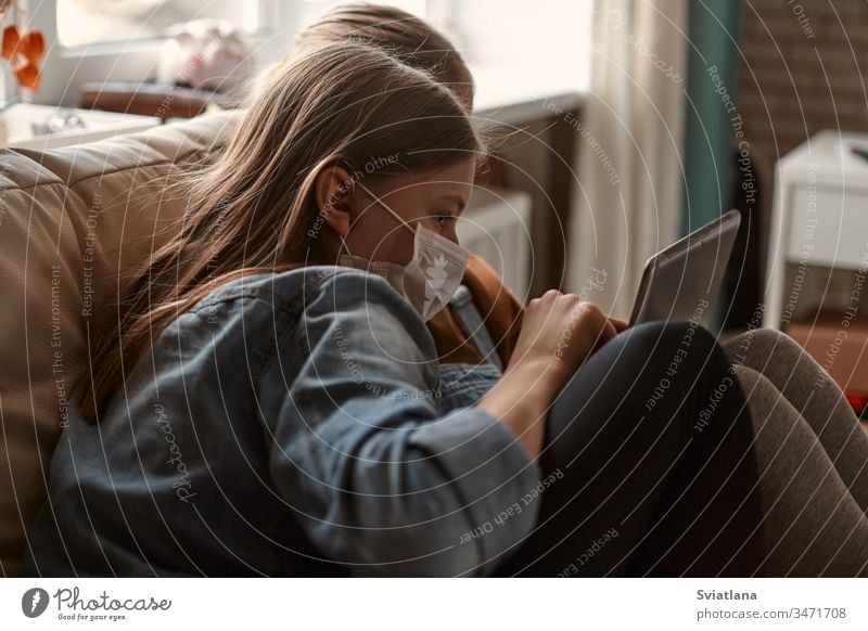 Two cute girls in medical masks are sitting on the sofa with a tablet. Schoolgirls do their homework. Distance learning during the COVID-19 quarantine.