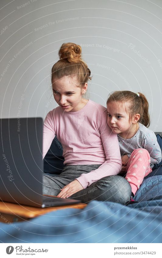 Teenage girl and her younger sister playing learning on laptop spending time together at home during COVID-19 quarantine. Girls sitting on bed in front of computer