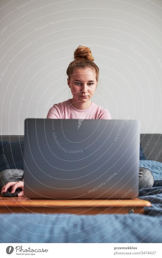 Teenage girl working on school tasks homework remotely on her laptop from home during COVID-19 quarantine. Online course lessons. Learning remotely. Girl sitting on bed in front of computer looking at screen