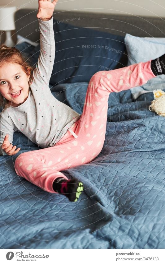 Little happy playful kid girl having fun making silly funny faces jumping on bed in bedroom in the morning at home playing childhood joy cute laughing smiling