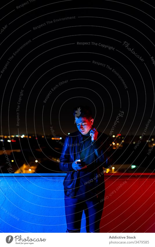 Young man using smartphone on illuminated balcony night colorful dark modern urban male red light blue light city device browsing street gadget evening