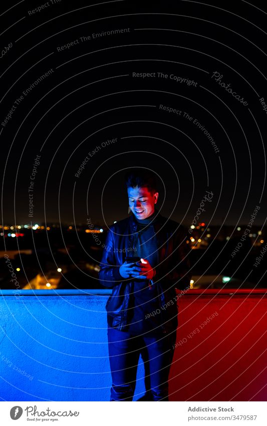 Young man using smartphone on illuminated balcony night colorful dark modern urban male red light blue light city device browsing street gadget evening