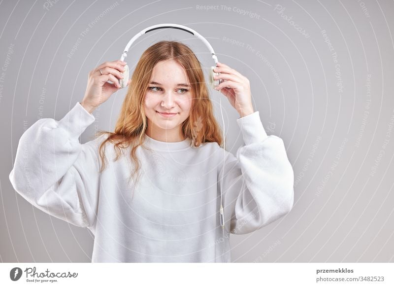 Young woman girl listening to music streaming content having fun watching video enjoying video chat talking with friends making gestures faces using smartphone earphones headphones standing over plain grey background