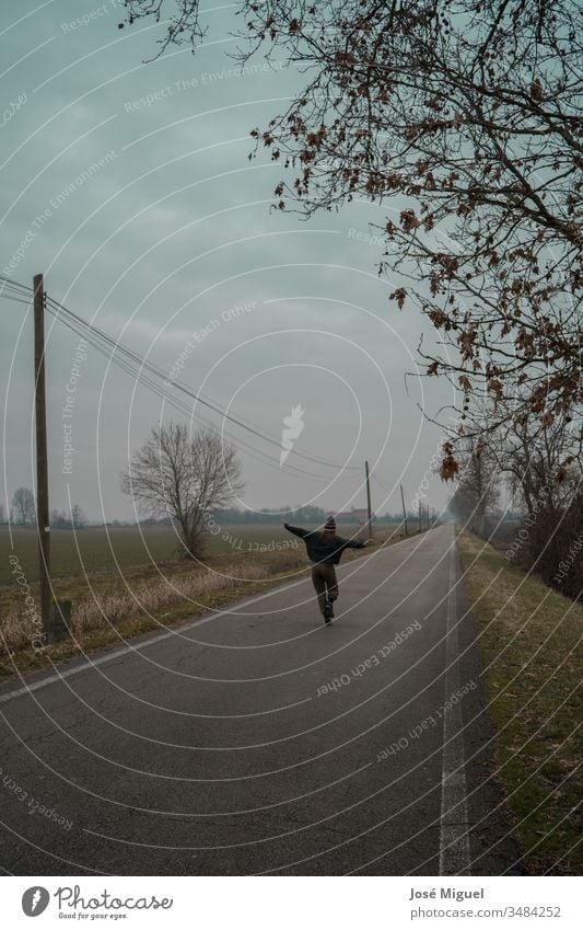 A girl walks down an asphalt road. The gray sky and blue background show an image in a farm field. The long highway has trees on the sides. landscape nature