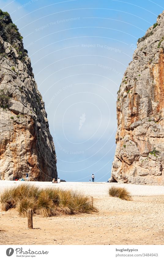 View over a mediterranean beach between two big rocks to the sky. Between the two rock faces, several small people can be seen sitting and walking. Beach Coast