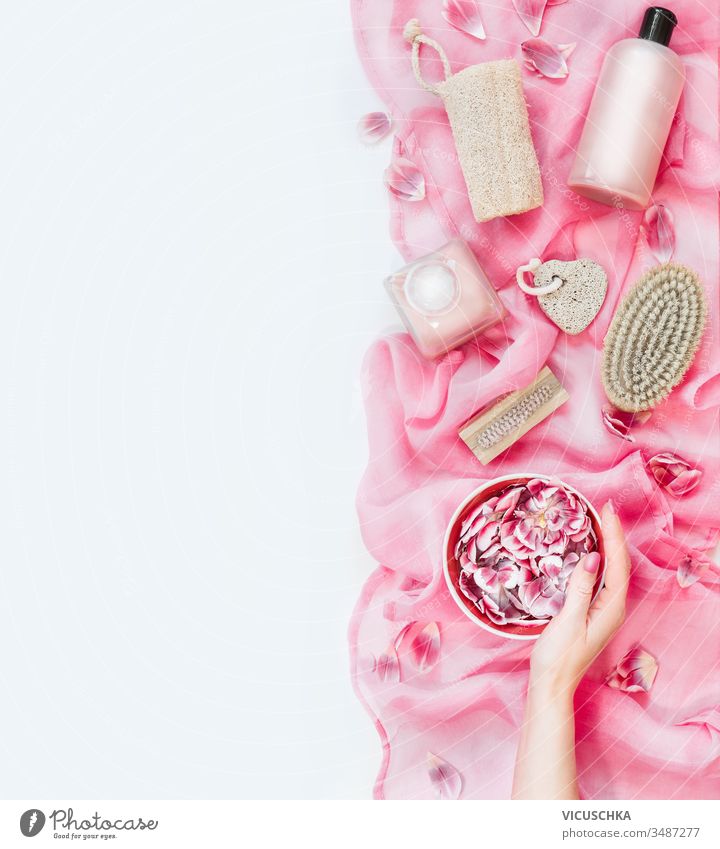 Female hand holding water bowl with flowers on pink towel with various eco friendly skin care and beauty tools: brush, sponge, pumice and cosmetic products on white background. Top view. Flat lay