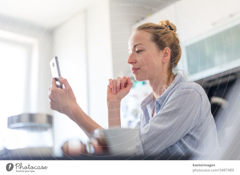 Young pleased woman indoors at home kitchen using social media apps on mobile phone for chatting and stying connected with her loved ones. Stay at home, social distancing lifestyle.