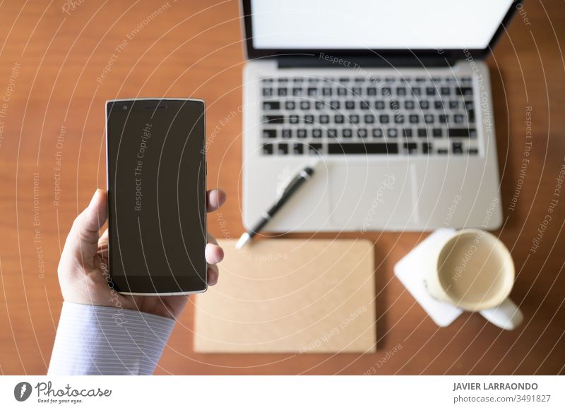 Young businessman holds in his hand a smart phone and in an unfocused background a wooden desk with a laptop, a notebook and a cup of coffee. working