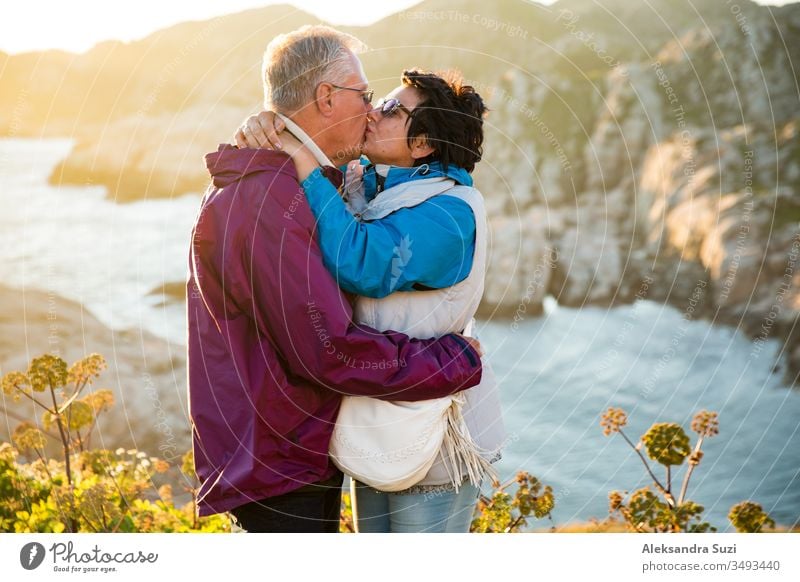 Loving mature couple traveling, standing on the top of rock, exploring. Active man and woman hugging and kissing, Happily smiling. Scenic view of mountains and sea on sunset. Norway, Lindesnes