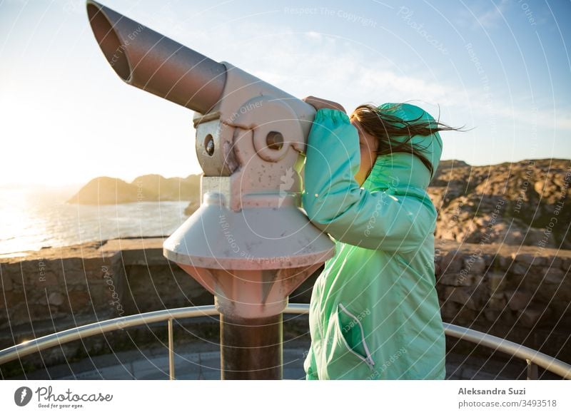 Teenage girl stands on rocky northern seashore, looking through touristic telescope, exploring the coastal rocks. Travel and enjoy adventure in Norway. Beautiful view of fjord and mountains in sunset