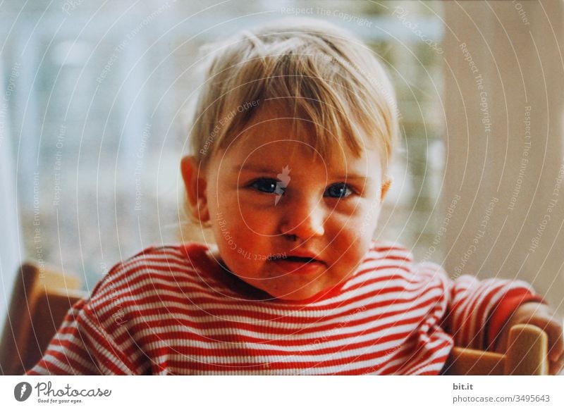 Cute, cute little girl in a striped shirt sits on a high chair and after dinner she smiles full, sly, cheeky, positive, happy and optimistically into the day.