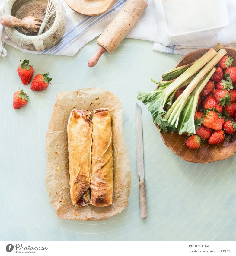 Strawberry rhubarb strudel cake on light mint kitchen table with knife and ingredients. Top view. Homemade seasonal baking strawberry top view homemade