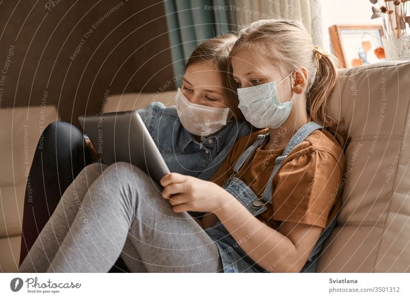 Two cute girls in medical masks are sitting on the sofa with a tablet. Schoolgirls do their homework. Distance learning during the COVID-19 quarantine.