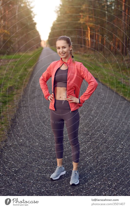 Full length front portrait of sporty young girl in training wear with grey pants and red jacket, standing and resting after jogging with her hands on hips, looking at camera and smiling