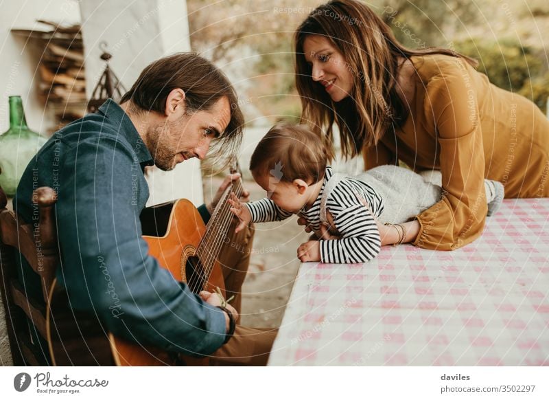 One year baby touching his father’s guitar while he plays at home courtyard. Mother is holding the baby. small calm garden casual style outdoors white lovely