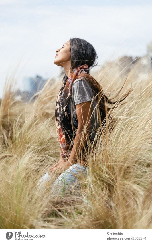 portrait of a young brunette woman with long hair wearing a scarf around her neck sitting on her knees in a wheat field with a calm expression smell wind feel