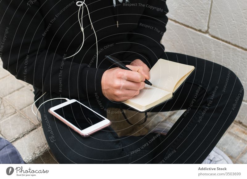 Crop student listening to music and writing in notebook man street write smartphone casual step male city notepad stair pen education sit study urban sound