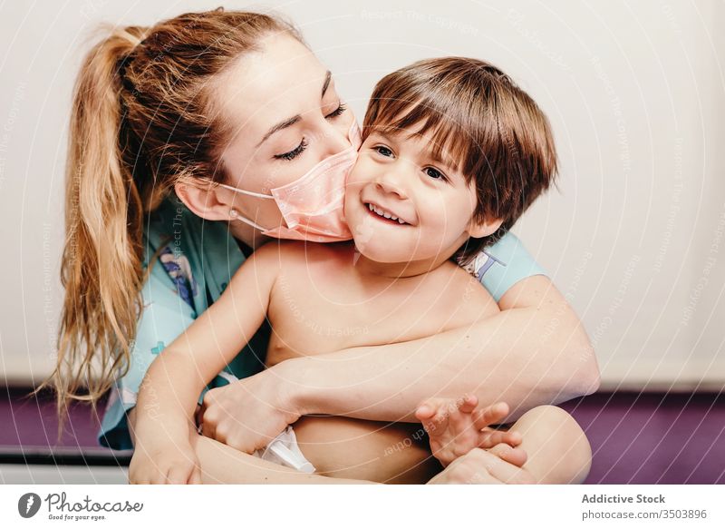 Happy woman in medical mask kissing kid happy patient clinic kind love hug prevent protect virus together cheerful young female cute little son child care smile