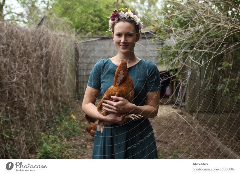 Young woman with a wreath of flowers in her hair stands in the chicken run holding a brown chicken in her arms Central perspective Shallow depth of field Day