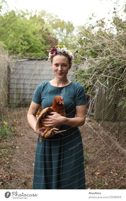 Young woman with a wreath of flowers in her hair stands in the chicken run holding a brown chicken in her arms Central perspective Shallow depth of field Day