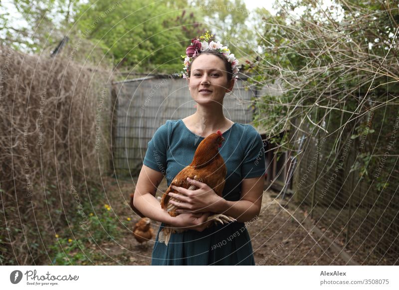 Young woman with a wreath of flowers in her hair stands in the chicken run holding a brown chicken in her arms Central perspective Shallow depth of field Day