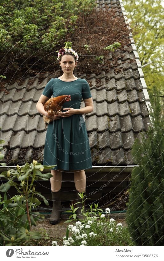 Young woman with a wreath of flowers in her hair stands in front of the summer house and holds a brown chicken in her arms Central perspective