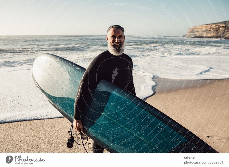 Smiling senior man carrying surfboard at beach looking at camera happy men vacation surfing adult smile grey hair lifestyle joy sport hobby portrait beard ocean