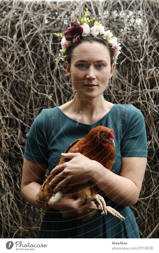 Young woman with a wreath of flowers in her hair stands in the chicken run holding a brown chicken in her arms Central perspective Shallow depth of field Day