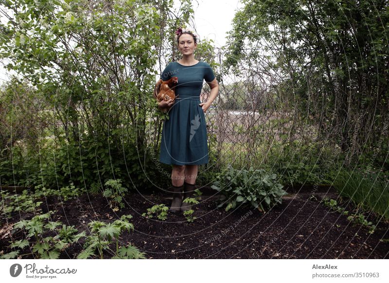 Young woman with wreath of flowers in her hair is standing in the garden holding a brown chicken in her arms Central perspective Shallow depth of field Day