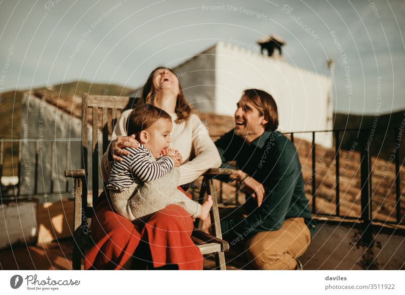 Parents laughing and having fun with their baby son sitting at the home terrace, with beautiful sunset light. real authentic alternative patio cheerful wife