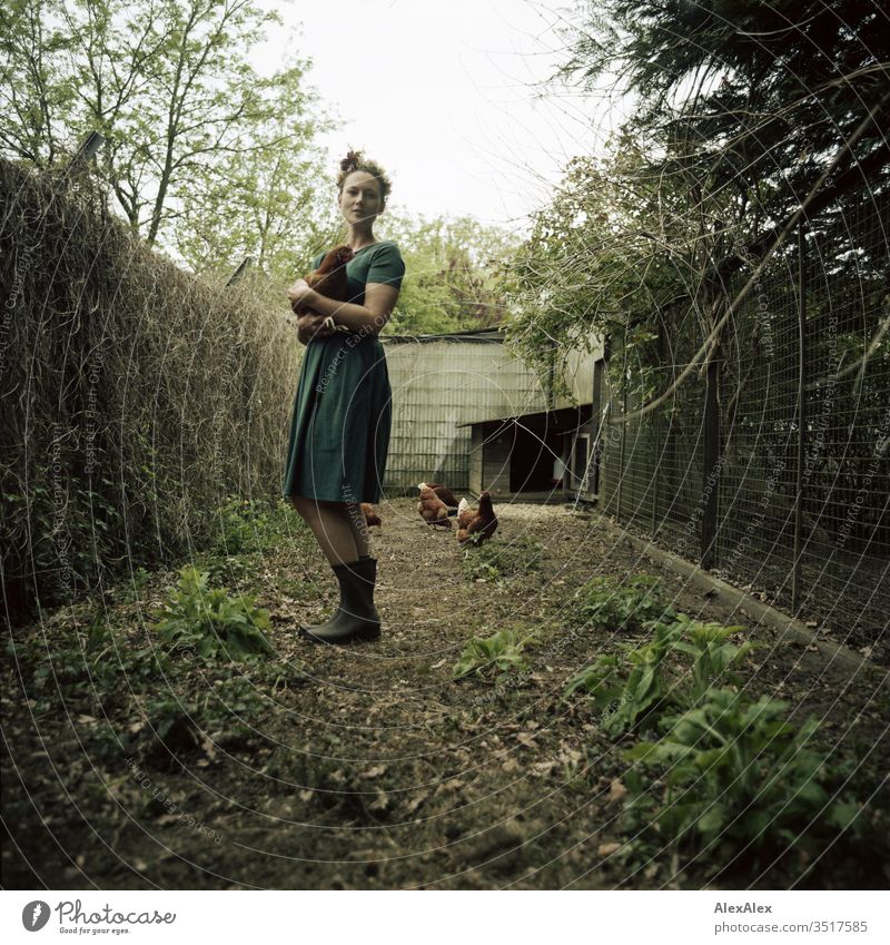 Young woman with a wreath of flowers in her hair stands in the chicken run holding a brown chicken in her arms Central perspective Shallow depth of field Day