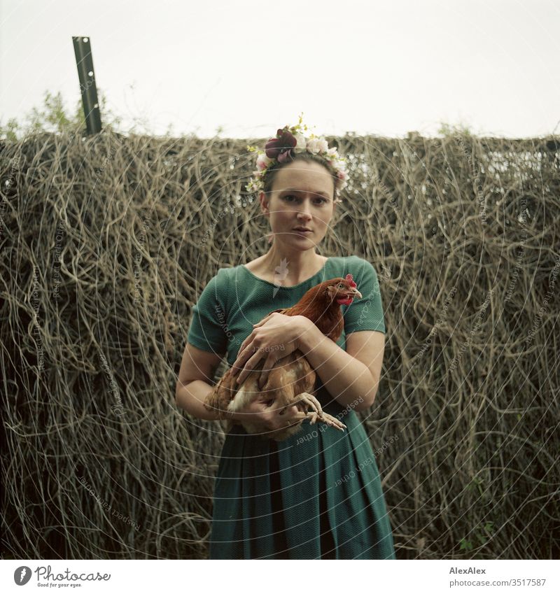 Young woman with wreath of flowers in her hair is standing in the garden holding a brown chicken in her arms Central perspective Shallow depth of field Day