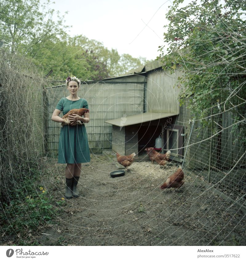 Young woman with a wreath of flowers in her hair stands in the chicken run holding a brown chicken in her arms Central perspective Shallow depth of field Day