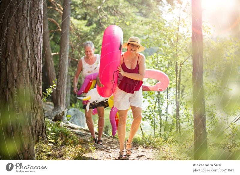 Elderly couple walking a forest path along the seashore holding giant inflatable flamingo and unicorn. Funny active pensioners enjoying summer vacation on the beach in Northern Europe