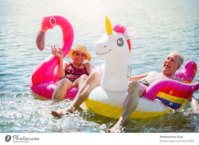 Elderly couple having fun on inflatable flamingo and unicorn. Funny active pensioners happy together enjoying summer vacation on the beach in Europe, laughing, playing the fool, splashing water.