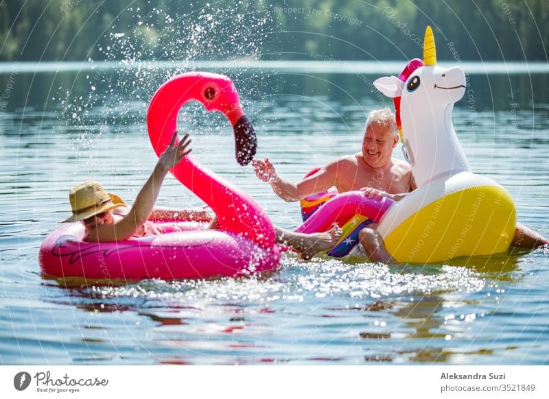 Elderly couple having fun on inflatable flamingo and unicorn. Funny active pensioners happy together enjoying summer vacation on the beach in Europe, laughing, playing the fool, splashing water.