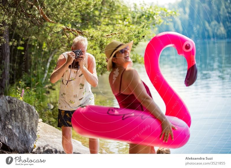 Elderly couple taking photos on vintage camera in forest along the seashore holding giant inflatable flamingo. Funny active pensioners enjoying summer vacation on the beach