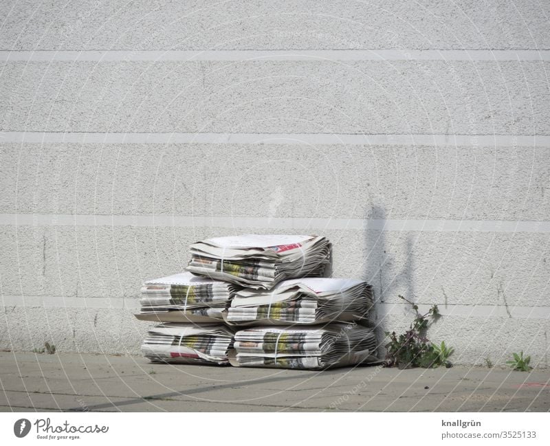 Five packs of newspapers stacked on the sidewalk in front of a white house wall, next to them some weeds growing out of a joint Newspaper Print media Stack