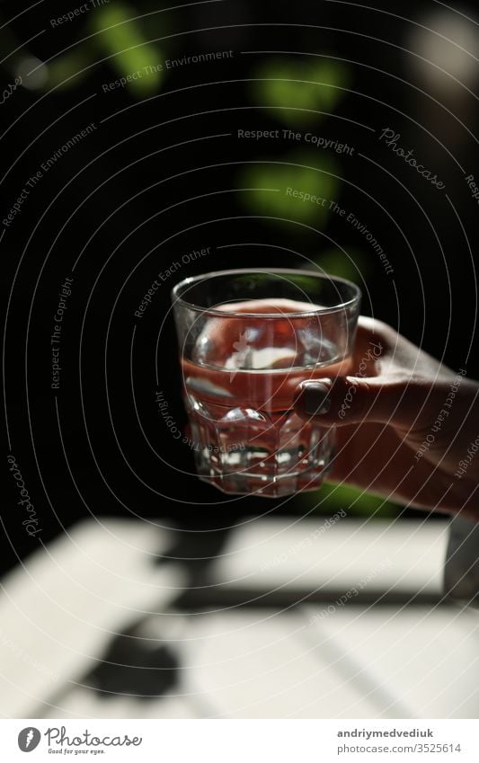 Woman hands isolated, holding a glass of water on a dark background with green leaves. the sun's rays fall on the glass. healthy morning drink natural life