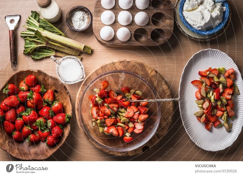 Fresh strawberries and rhubarb on wooden table background with ingredients for tasty seasonal cooking or baking. Top view. Healthy clean food. Paleo dieting. Home cuisine. Garden fruits eating