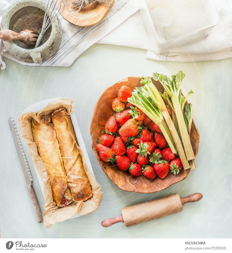 Strawberry rhubarb wrap cake on kitchen table  with ingredients. Top view. Summer baking strawberry background bowls top view summer seasonal products