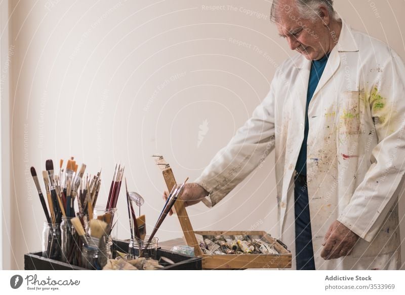 Senior artist standing in home workshop and choosing paints man aged set paintbrush choose concentrate studio hobby gallery equipment modern table design